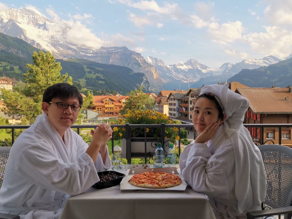 Couple enjoying a meal on a hotel balcony in the Swiss Alps, with panoramic mountain views, traditional chalet rooftops, and lush green valleys in the background.