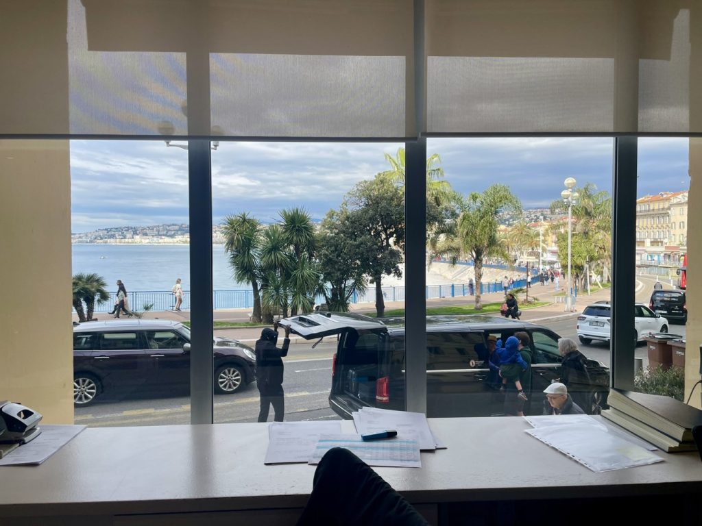 Hotel Suisse Nice Review: Iconic Sea Views on the French Riviera 6 View of Baie des Anges and Promenade des Anglais from Hôtel Suisse Nice window with palm trees and people walking by.