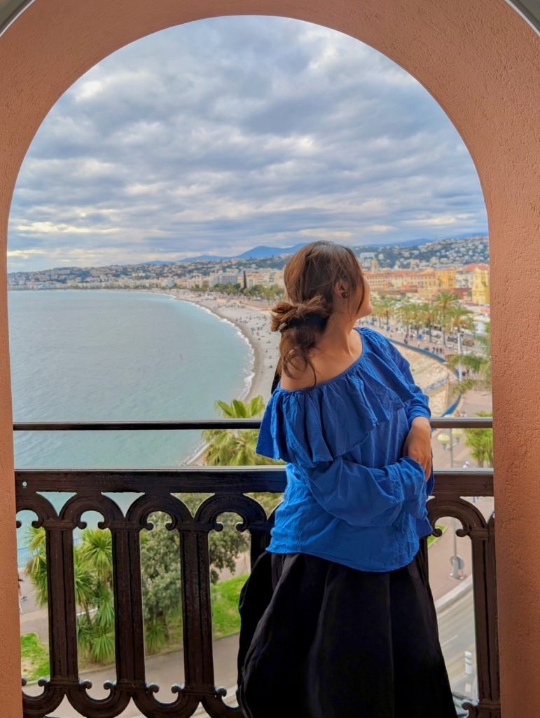 Hotel Suisse Nice Review: Iconic Sea Views on the French Riviera 16 Guest in blue blouse enjoying panoramic sea view of Baie des Anges from Hôtel Suisse Nice arched balcony.