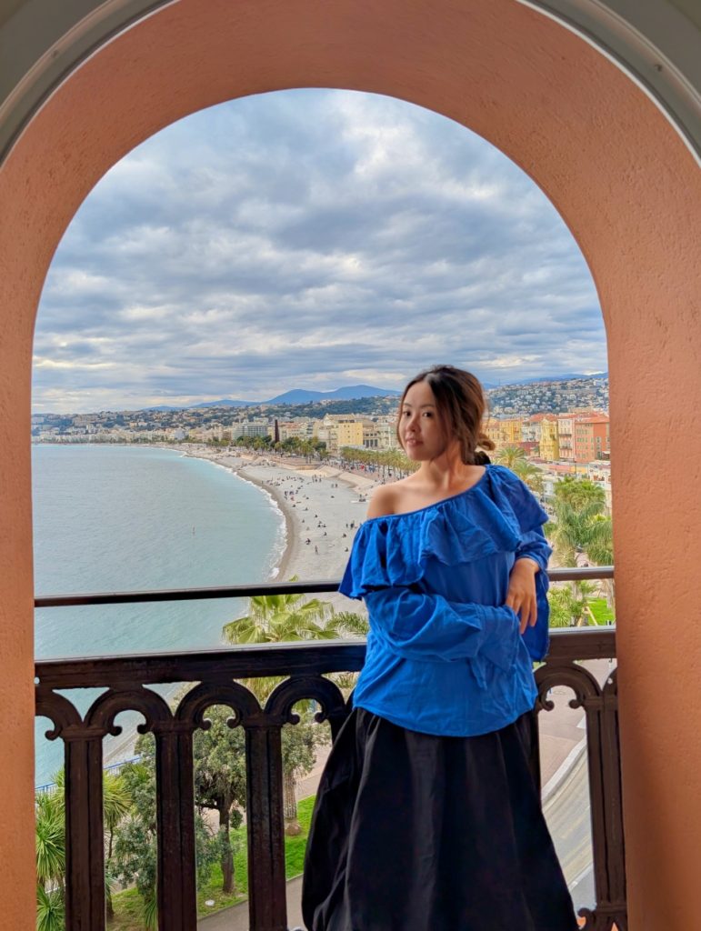 Hotel Suisse Nice Review: Iconic Sea Views on the French Riviera 17 Guest posing on arched balcony at Hôtel Suisse Nice with sweeping view of Promenade des Anglais and Mediterranean Sea.