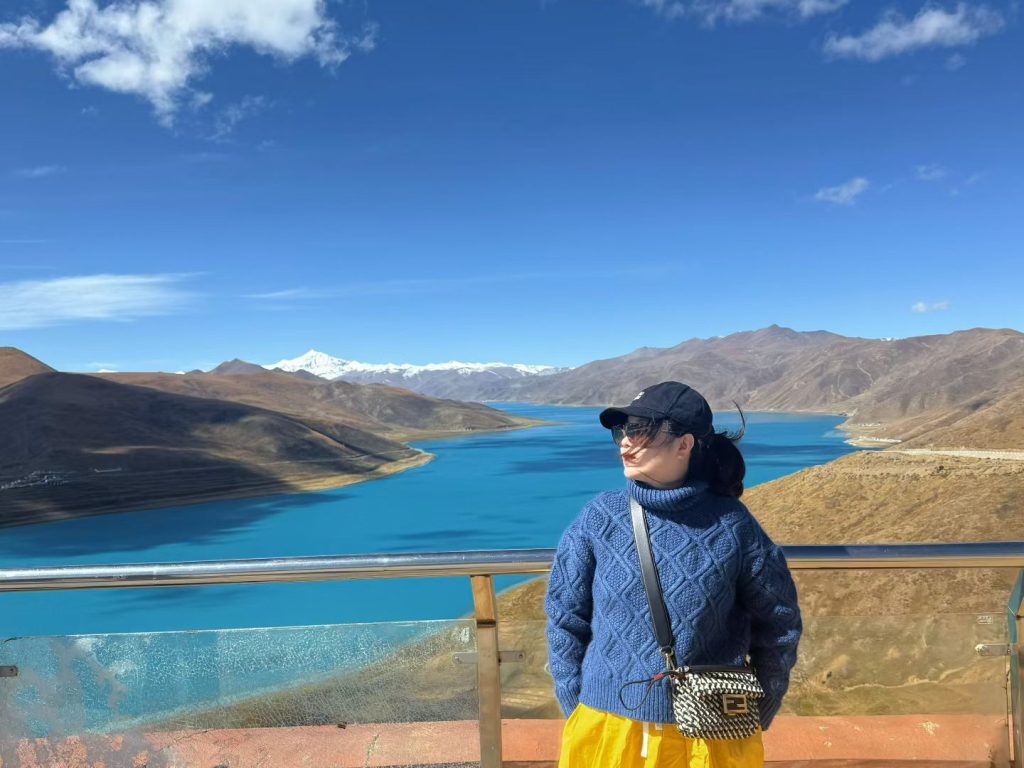 Nicole standing by Yamdrok Lake, wind blowing her hair as she gazes across the turquoise water