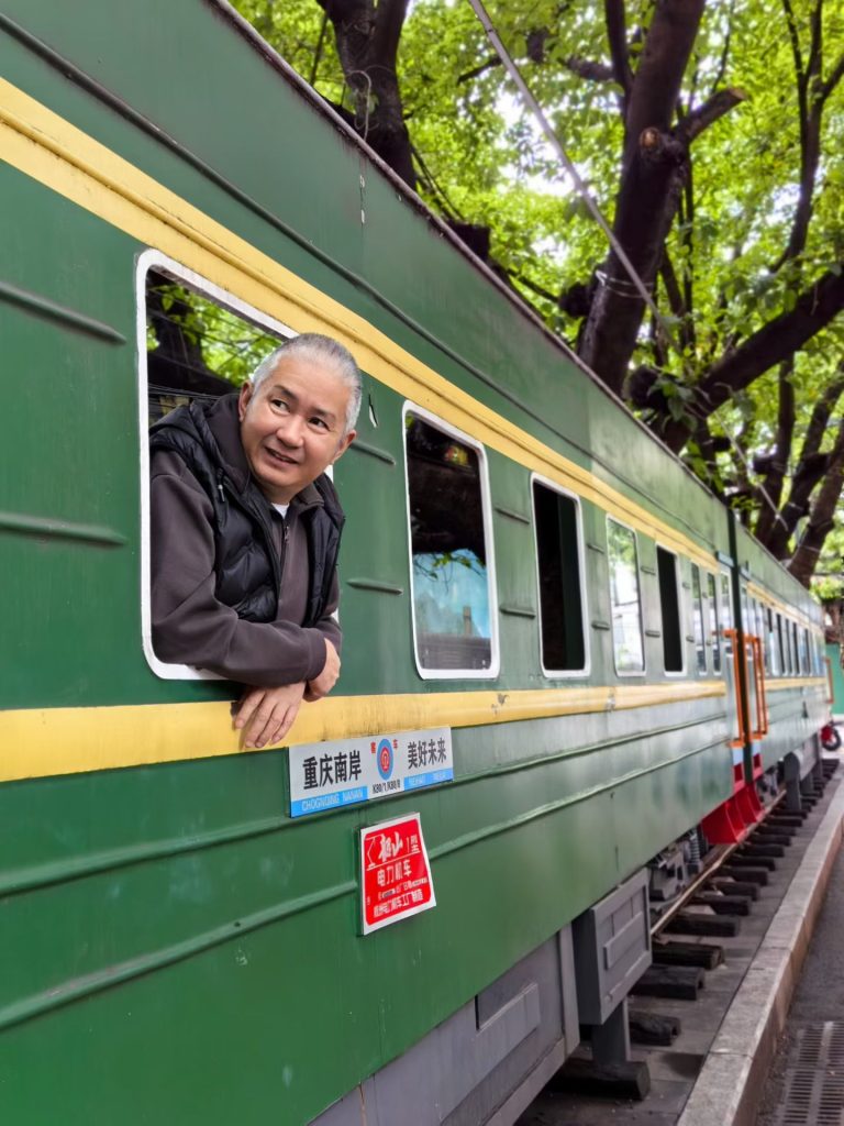 Man leaning out of the window of a vintage green train carriage under shady trees in Super 80 Street.