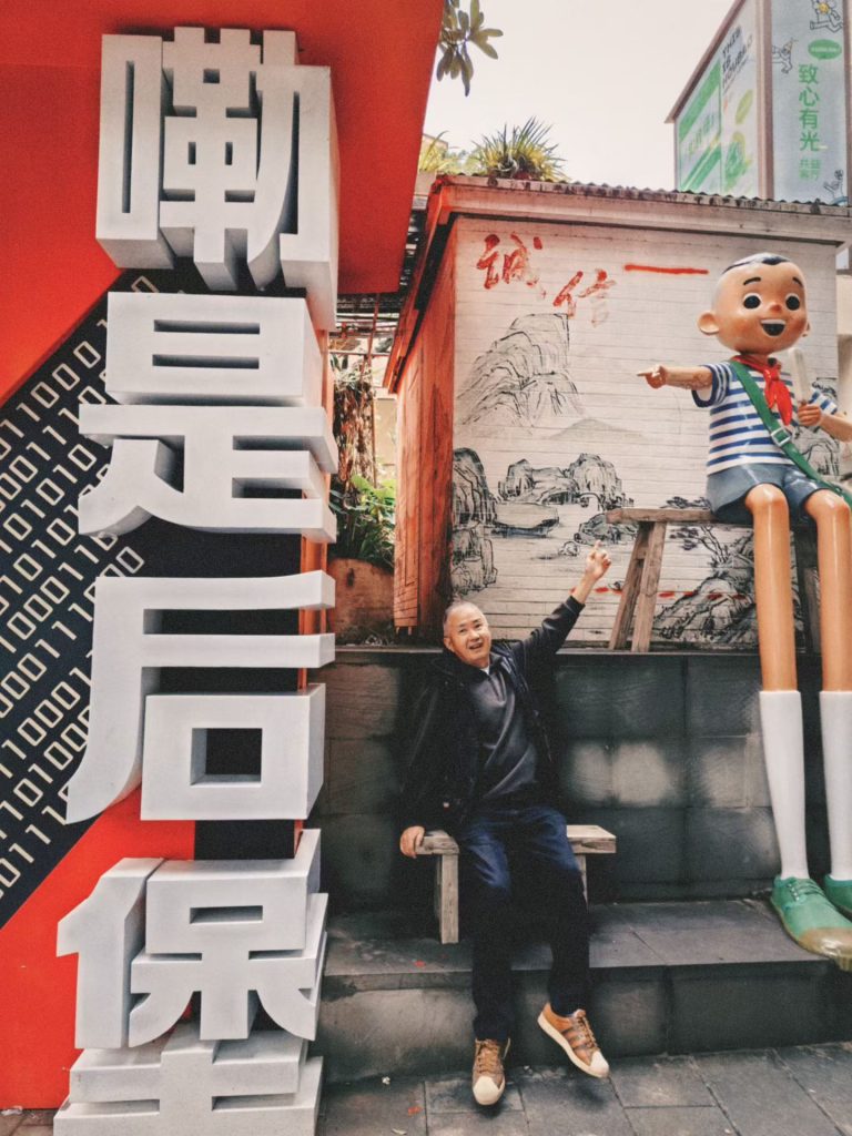 Man sitting next to a tall cartoon boy sculpture with long legs on Super 80 Street in Chongqing.