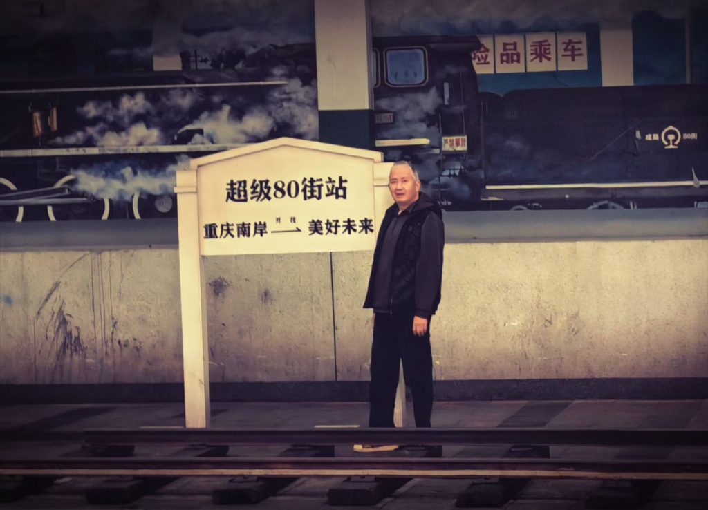 Man standing beside a sign reading “Super 80 Street Station” with a mural of an old train in the background.