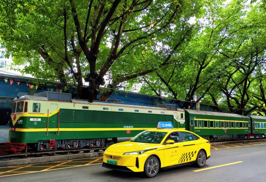 Retro green train carriage parked along the street with a yellow Chongqing taxi passing in front.
