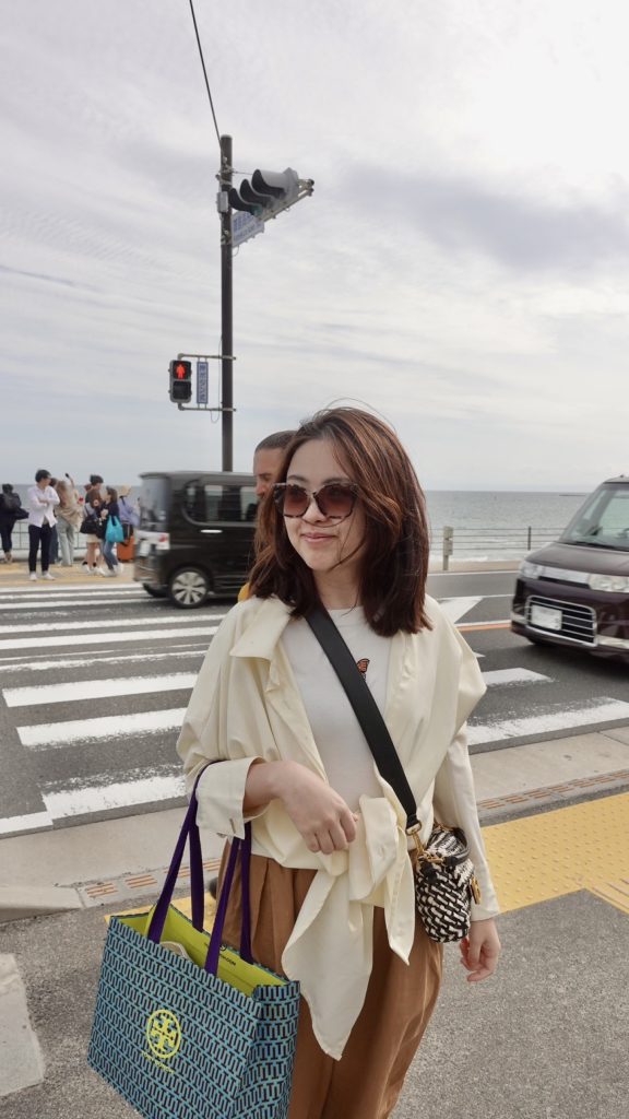 Kamakurakokomae Station: Visiting the Iconic Slam Dunk Crossing by the Sea 7 A woman smiling near the Kamakurakokomae Station crossing as cars pass by under clear blue skies