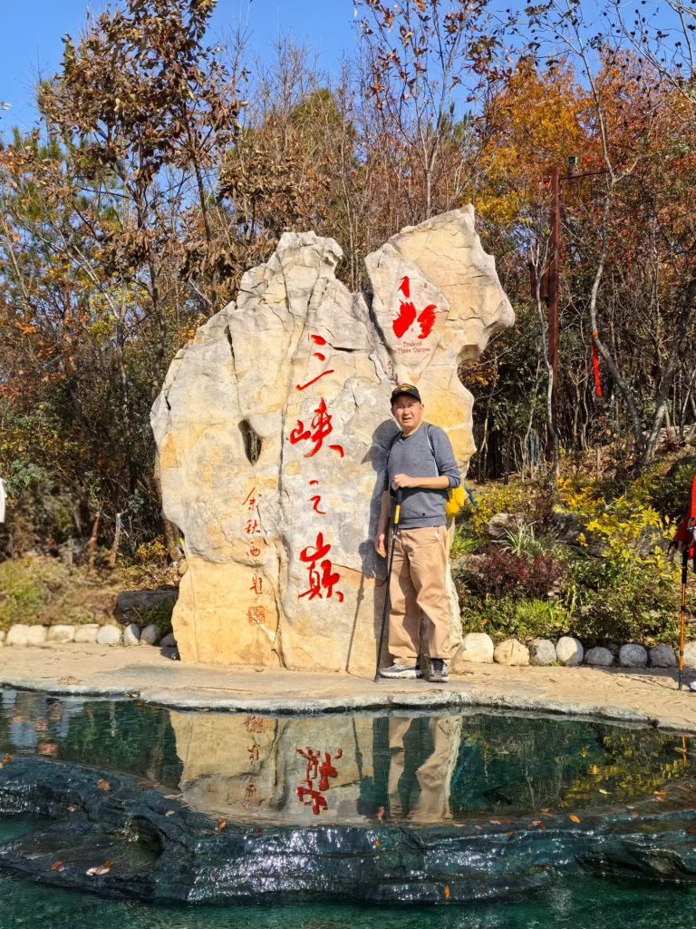 Hiking the Three Gorges Summit in Wushan: Stunning Views & Autumn Colors 13 Visitor standing beside the Three Gorges Summit stone marker with a clear reflection in the pool of water below.