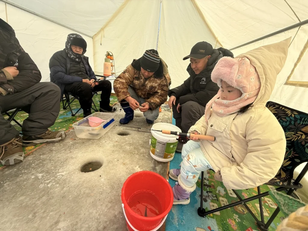 Child holding a small ice fishing rod inside the tent while adults prepare bait nearby