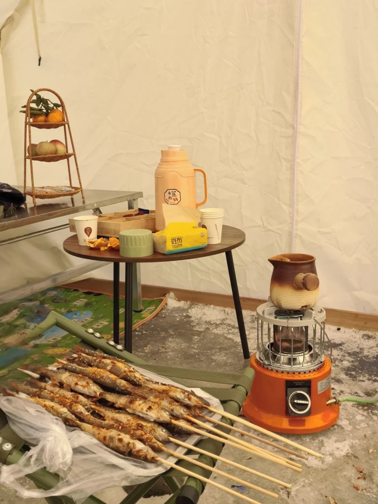 Ice fishing tent setup with heater, kettle, snacks, and grilled fish skewers in the foreground