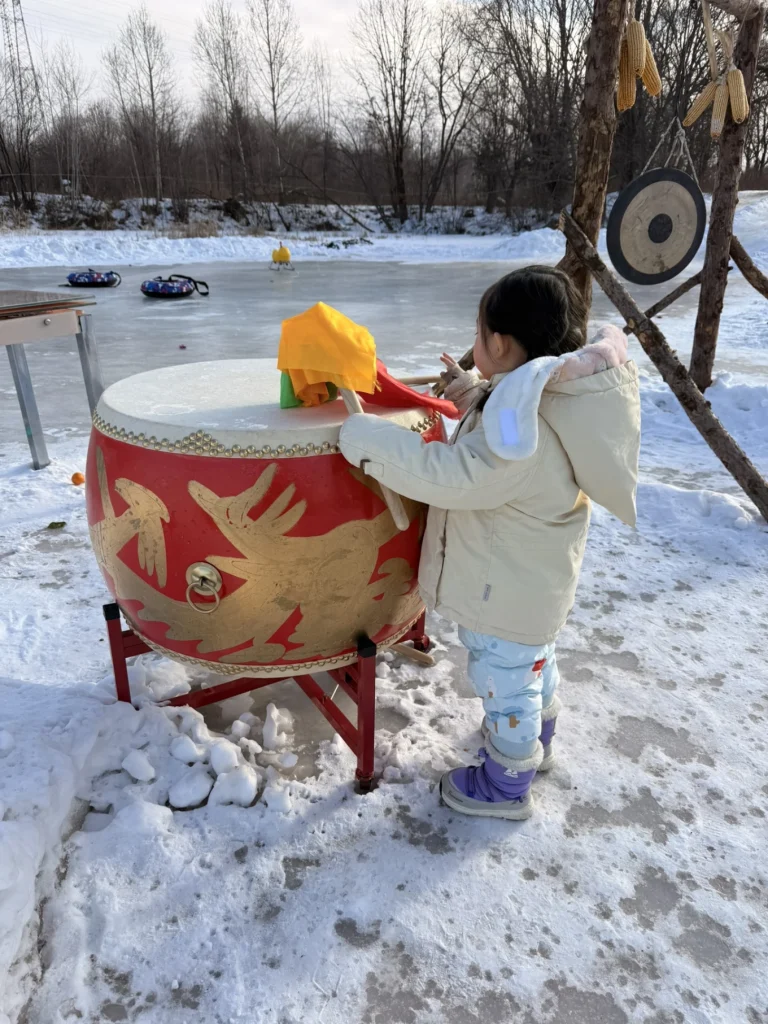 Child standing by a large red drum on snow near a target board at a winter activity area