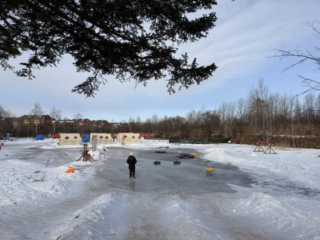 Wide view of frozen river activity area with tents, tubes, and snowbanks under tree branches