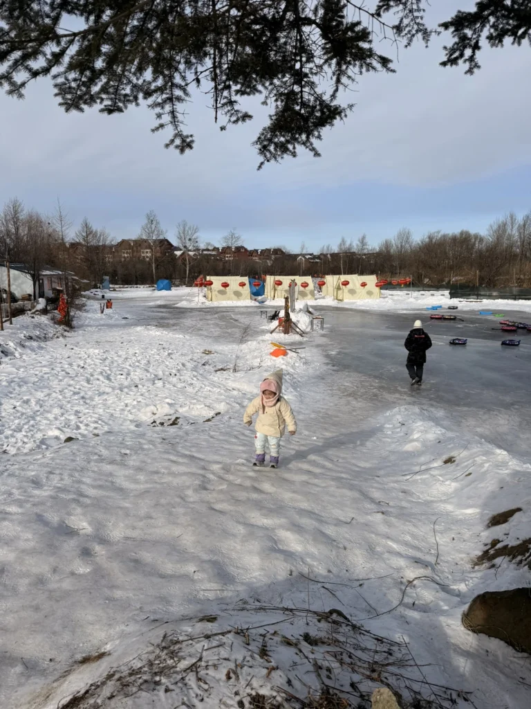Child walking uphill on snowy ground beside a frozen river with tents and tubes in the distance