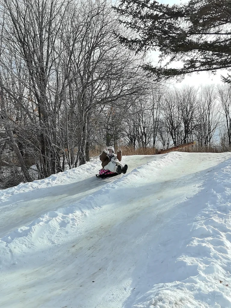Child riding a snow tube down a steep icy slope lined with snowbanks and bare trees