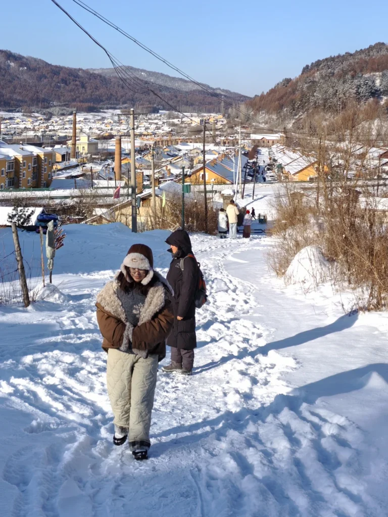 Hengdaohezi 1-Day Trip: Snowy Railway Town Near Harbin Travel Guide 17 Walking along a snowy hillside path in Hengdaohezi, overlooking the historic railway town and surrounding winter landscape.