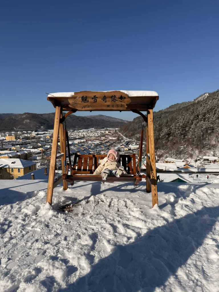 Child sitting on a wooden swing at a snowy viewpoint overlooking Hengdaohezi town and surrounding mountains in winter.