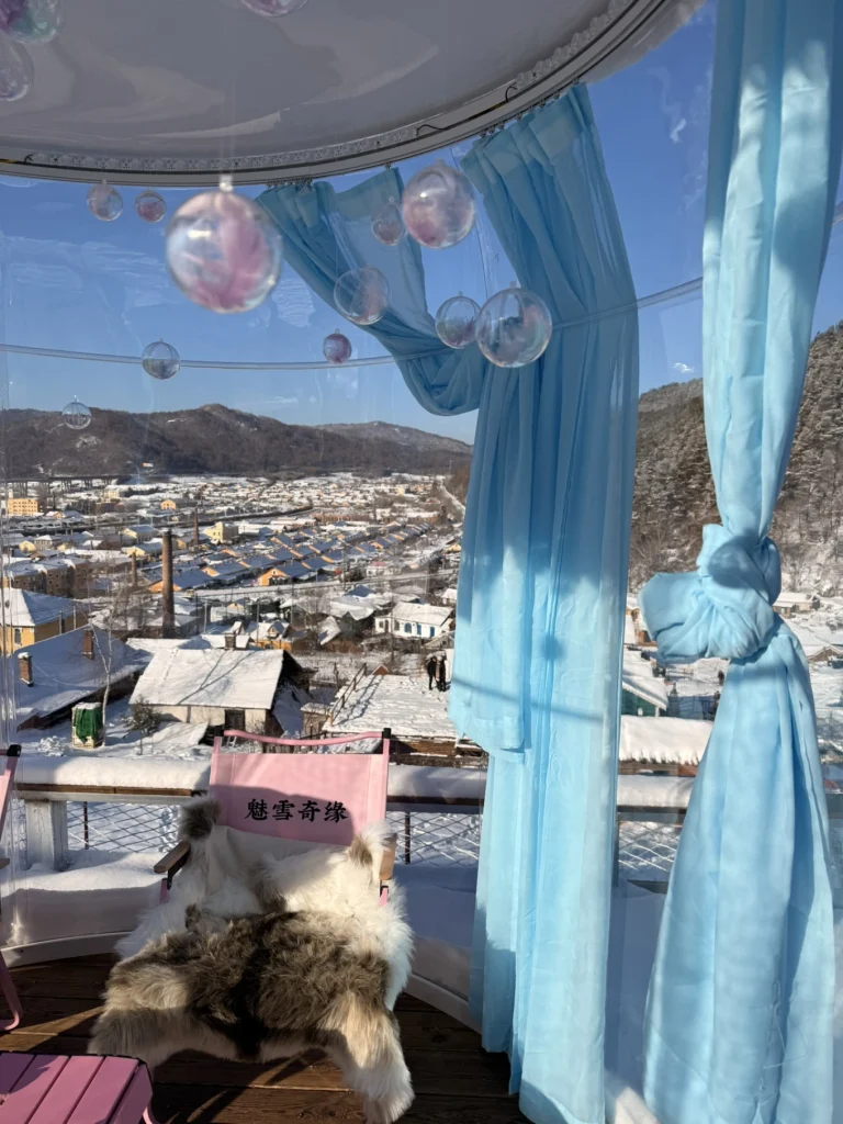 Interior of a transparent bubble tent café on a hillside in Hengdaohezi, with blue curtains and winter town views outside.