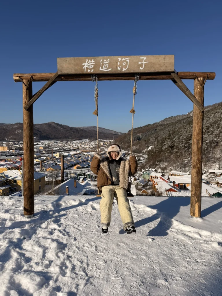 Me sitting on a wooden swing at a snowy viewpoint in Hengdaohezi, with the town and mountains in the background.