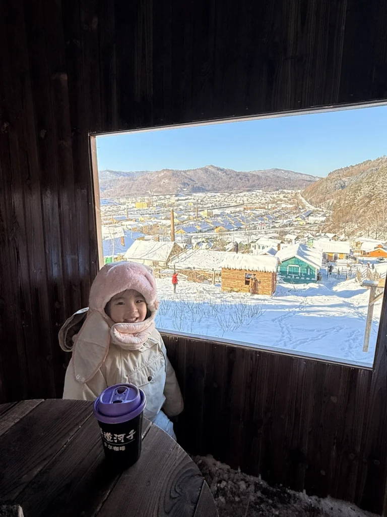 Child sitting inside a wooden viewing hut in Hengdaohezi, looking out over the snowy town through a large window.