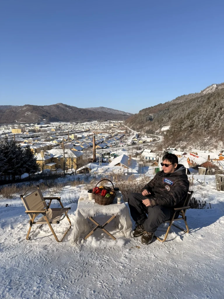 Winter hillside picnic spot overlooking Hengdaohezi town, with snow-covered rooftops and mountains in northeastern China.