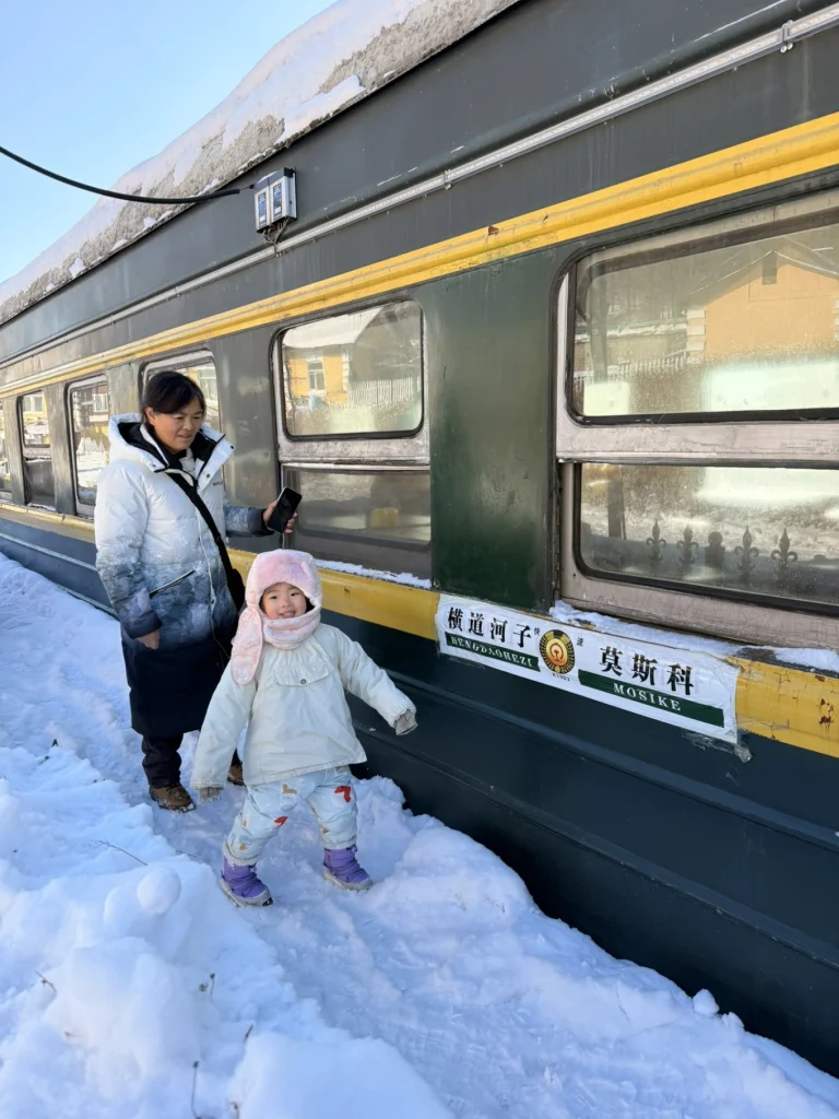 Hengdaohezi 1-Day Trip: Snowy Railway Town Near Harbin Travel Guide 26 Child and adult standing beside a historic green train carriage at Hengdaohezi Railway Town, Harbin, surrounded by deep winter snow.