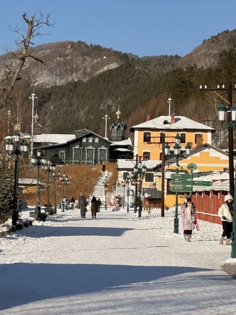 Snow-covered pedestrian street in Hengdaohezi, with historic Russian-style buildings, vintage street lamps, and forested hills in the background.