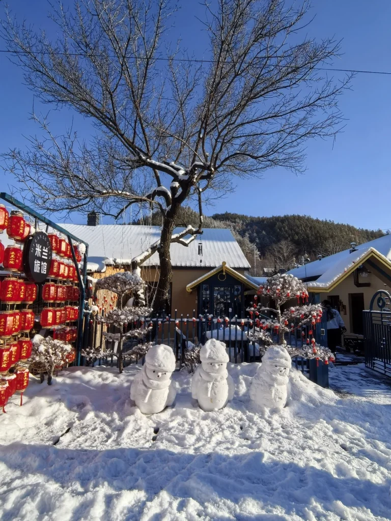 Hengdaohezi 1-Day Trip: Snowy Railway Town Near Harbin Travel Guide 7 Snowmen in front of a decorated guesthouse in Hengdaohezi during winter, with red lanterns, snowy rooftops, and forested hills behind.