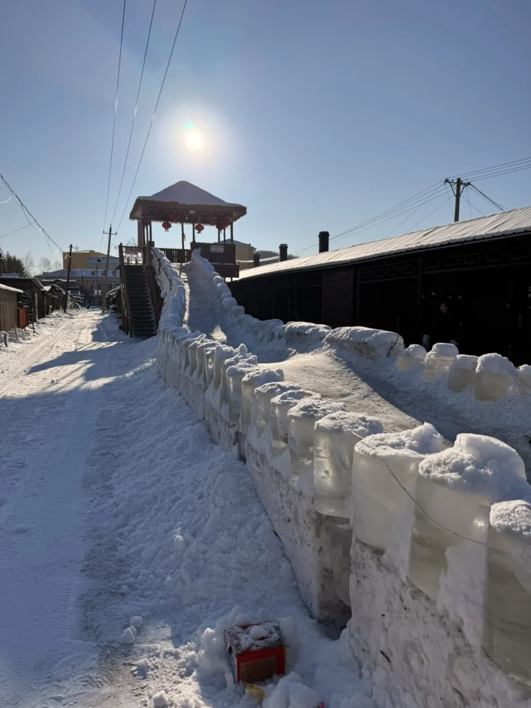 Hengdaohezi 1-Day Trip: Snowy Railway Town Near Harbin Travel Guide 15 Ice slide built along a snowy street in Hengdaohezi during winter, with sunlight, wooden houses, and clear blue sky.
