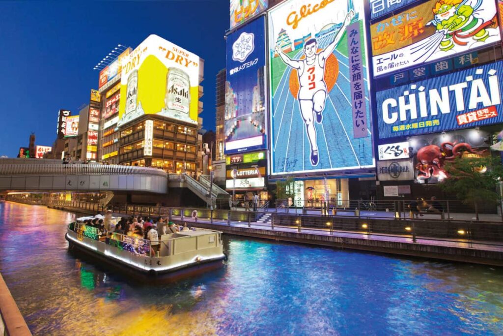 Dotonbori at night in Osaka, with bright neon billboards, the Glico Running Man sign, and a sightseeing boat cruising along the canal.