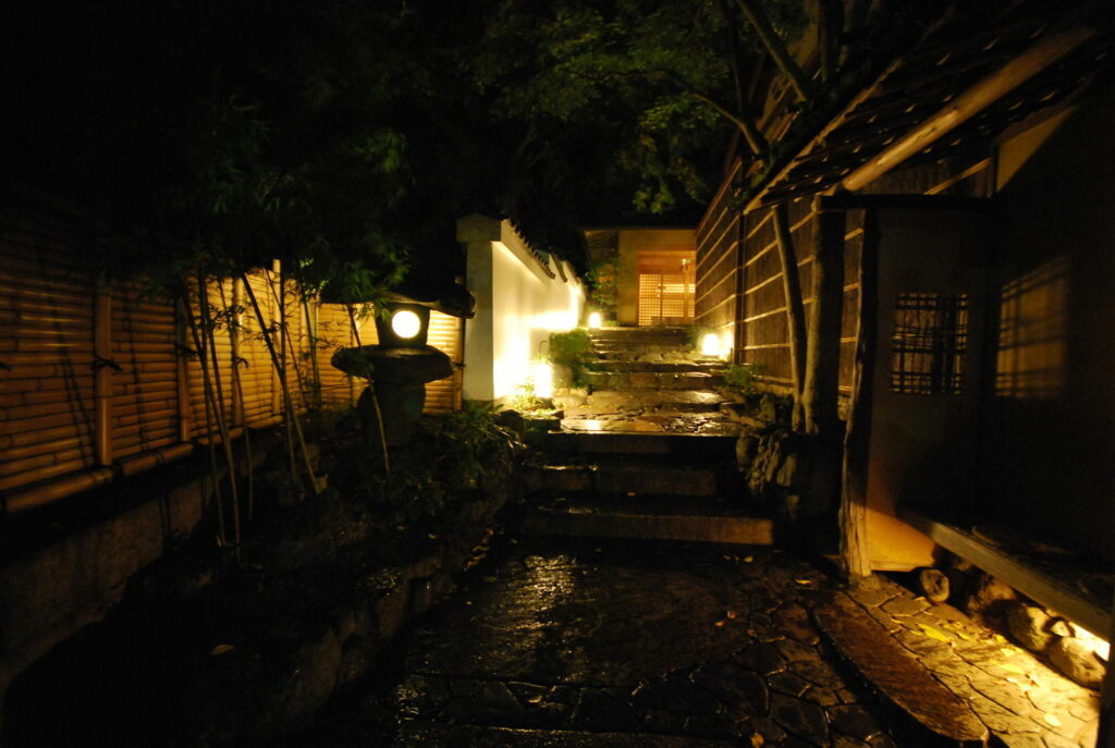 Gion Hatanaka ryokan entrance at night in Kyoto with illuminated stone pathway, bamboo fence, and traditional Japanese lantern lighting