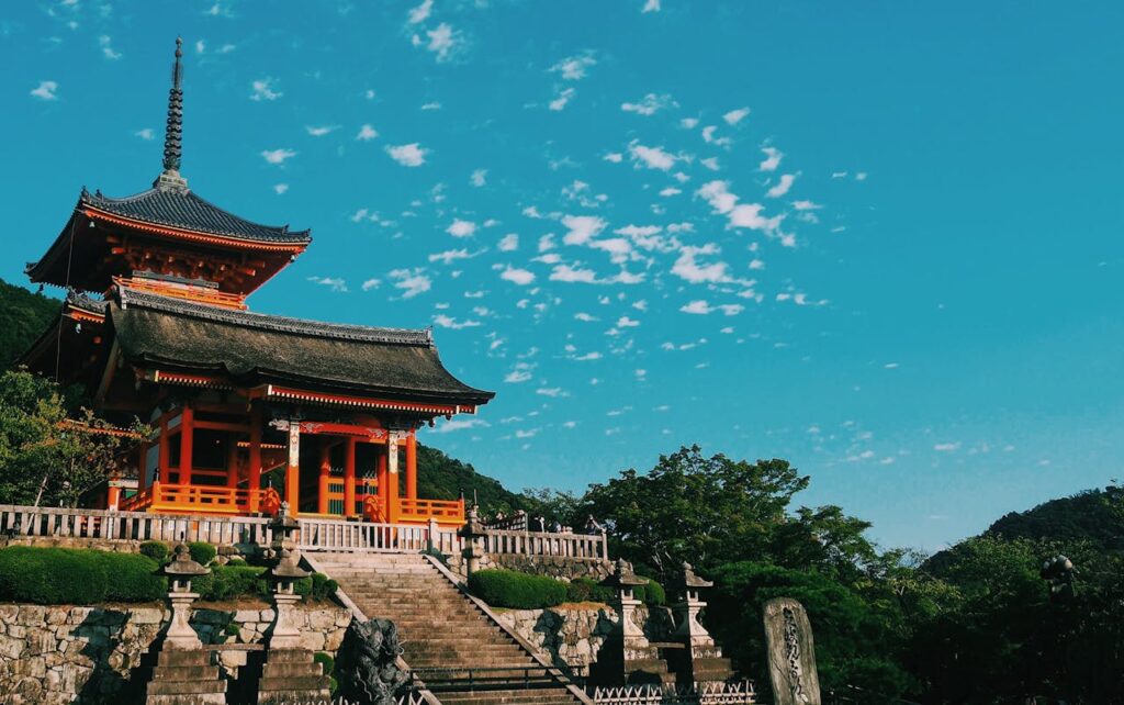 Traditional Japanese temple with red wooden pillars and pagoda-style roof overlooking stone steps and greenery under a bright blue sky