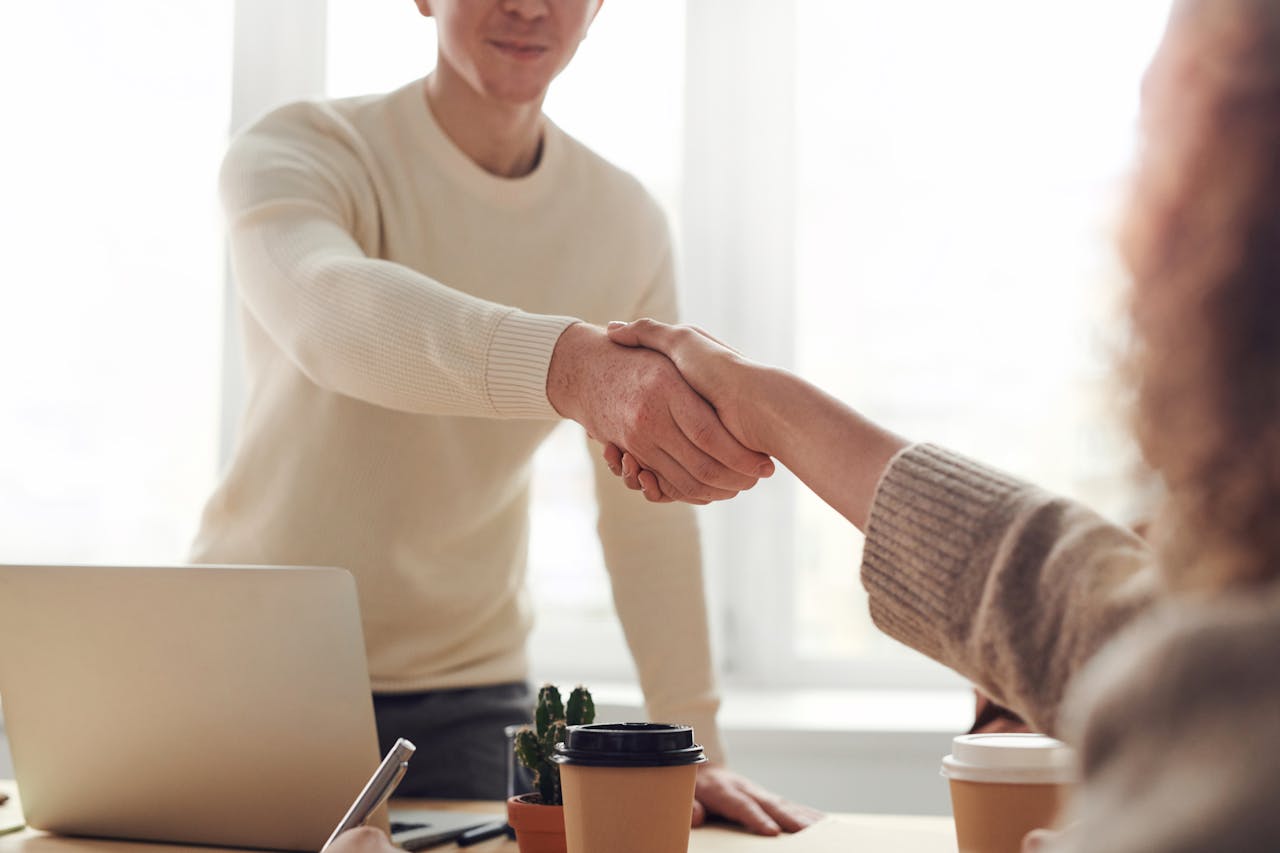 Partner With ShinyVisa 14 Business partnership handshake across a desk with laptop and coffee cups in a bright office setting
