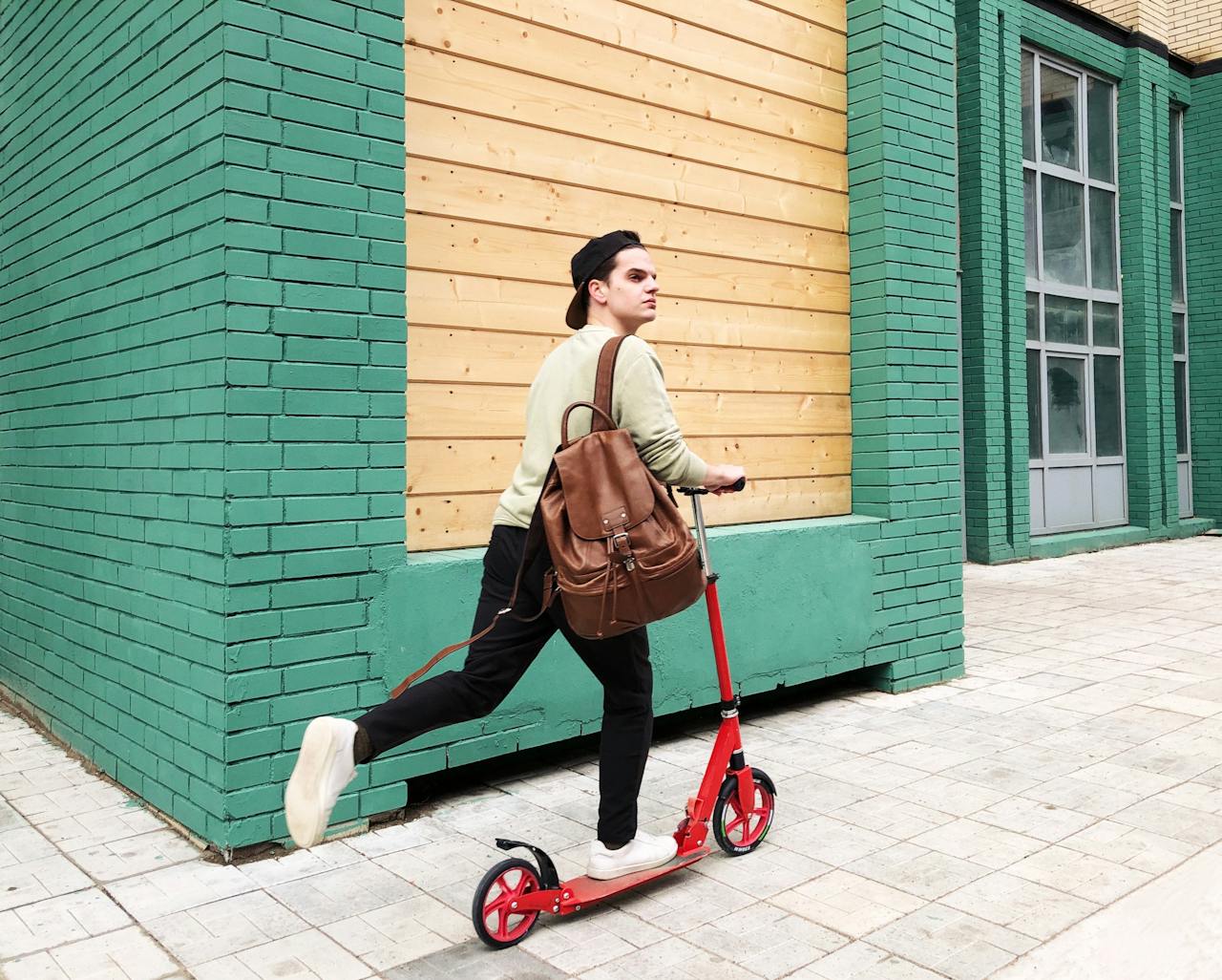 Young man riding a red kick scooter on a city sidewalk beside a teal brick building, wearing a backpack and casual clothes.