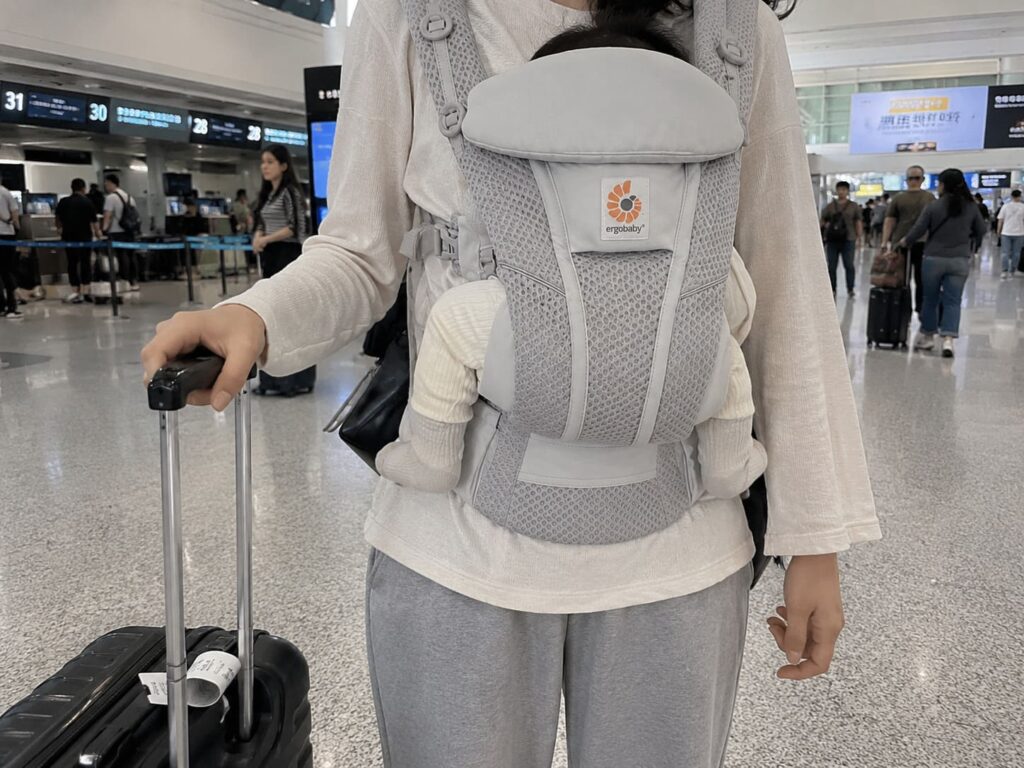 Chinese mother using a breathable Ergobaby baby carrier while walking through an airport terminal with luggage during family travel