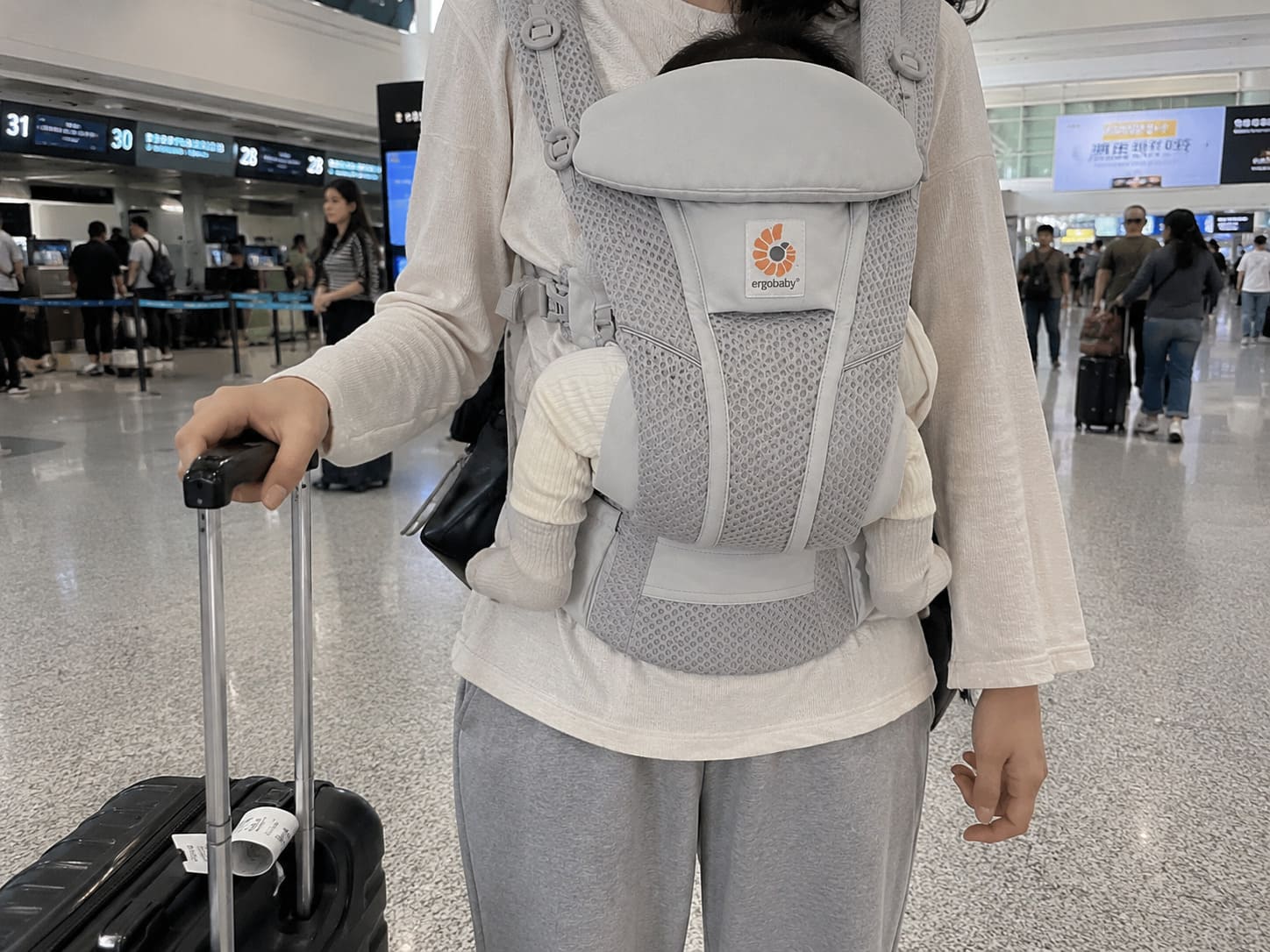 Chinese mother using a breathable Ergobaby baby carrier while walking through an airport terminal with luggage during family travel