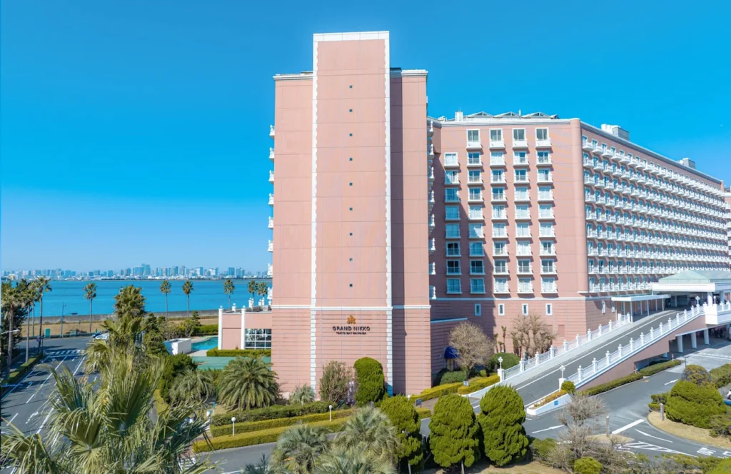 Exterior view of Grand Nikko Tokyo Bay Maihama overlooking Tokyo Bay, featuring the hotel’s pink facade, palm trees, and resort-style setting near Tokyo DisneySea.