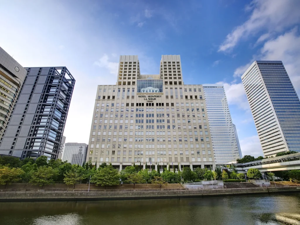 Exterior view of Hotel Monterey La Soeur Osaka beside the river, surrounded by modern skyscrapers under a bright blue sky in central Osaka.