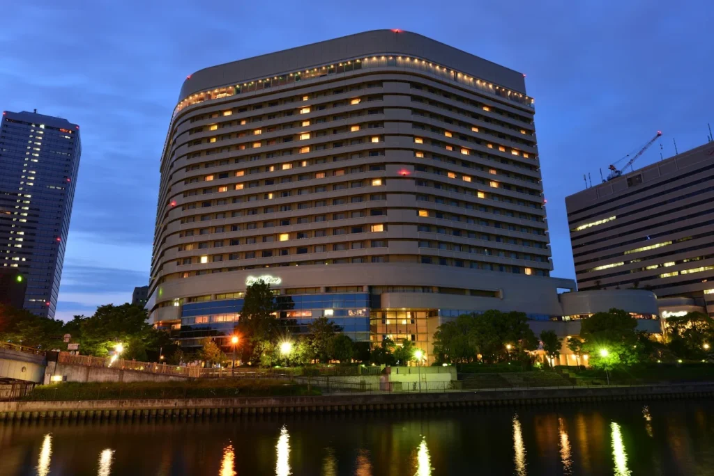 Exterior night view of Hotel New Otani Osaka beside the river, with warm lights glowing from the hotel windows and reflections shimmering on the water under a blue evening sky.