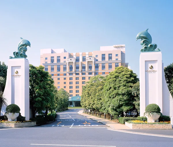 Entrance view of Hotel Okura Tokyo Bay framed by elegant white pillars topped with dolphin sculptures, leading toward the luxury hotel surrounded by lush greenery.