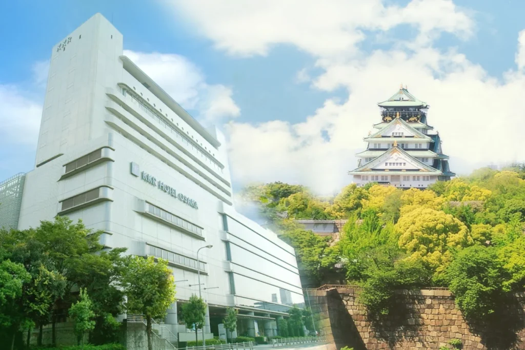 Exterior view of KKR Hotel Osaka beside Osaka Castle, showing the hotel building and the historic castle surrounded by lush green trees under a bright blue sky.