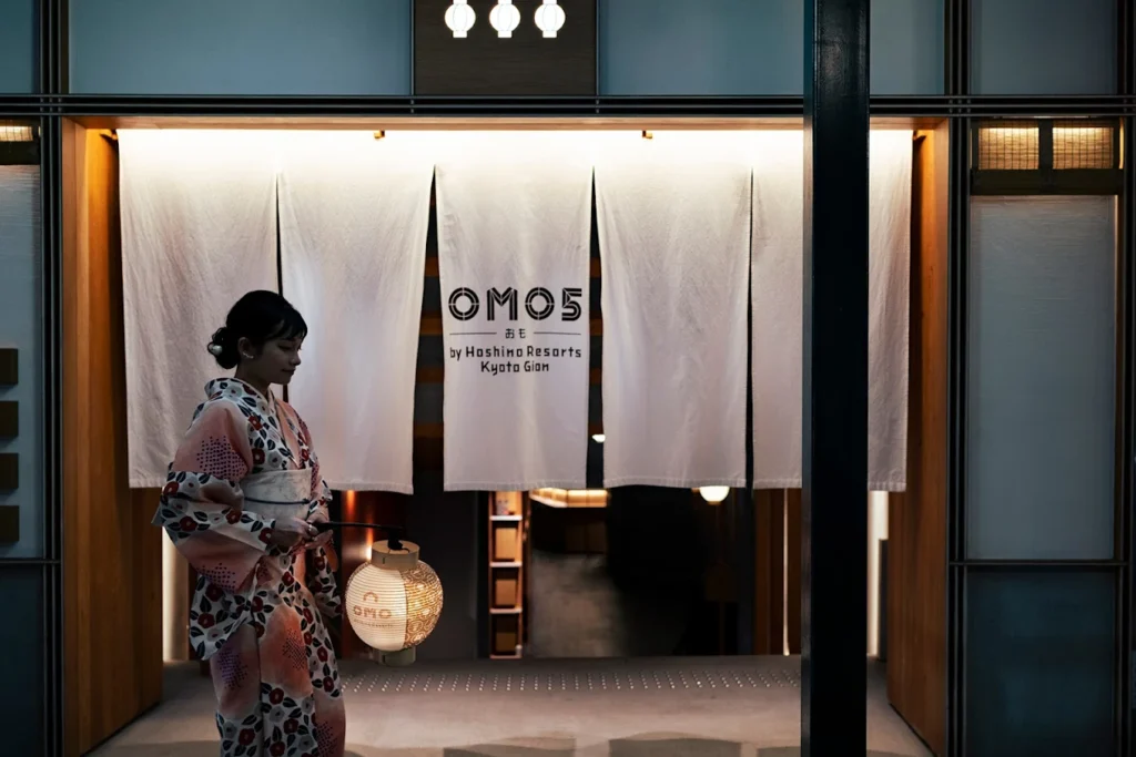 Entrance of OMO5 Kyoto Gion by Hoshino Resorts with a guest in a kimono walking past traditional noren curtains and warm ambient lighting in Kyoto.