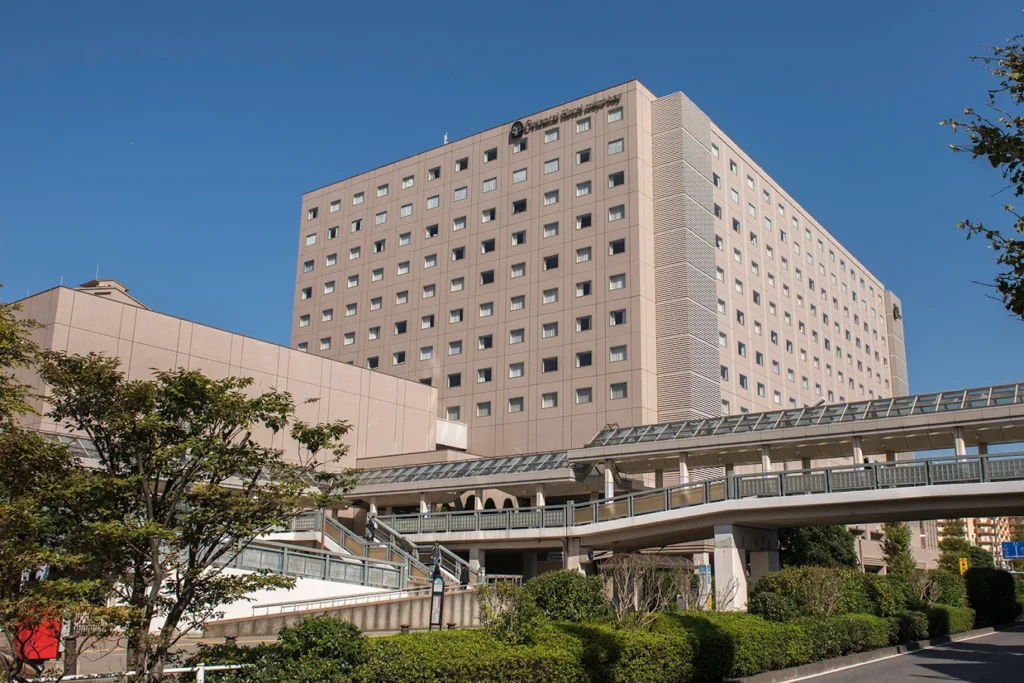 Exterior view of Oriental Hotel Tokyo Bay connected to Shin-Urayasu Station via elevated walkways, featuring a modern hotel facade under a clear blue sky.