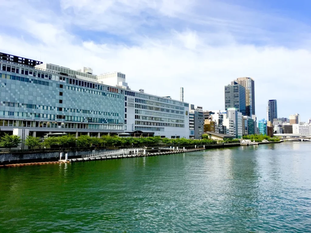 Riverside view of Osaka Castle Hotel and surrounding Osaka city skyline under a bright blue sky, with modern buildings lining the calm riverfront.