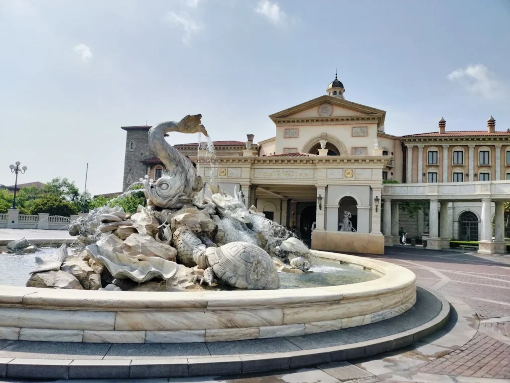 Exterior fountain entrance of Tokyo DisneySea Hotel MiraCosta on a sunny day, featuring an elaborate sea-themed sculpture with fish, turtles, and shell details in front of the Mediterranean-inspired luxury Disney hotel.