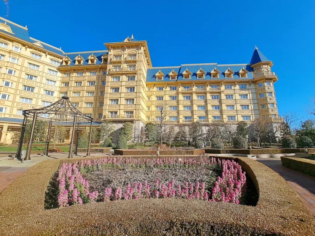 Exterior view of Tokyo Disneyland Hotel featuring elegant Victorian-inspired architecture, landscaped gardens, and colorful flower beds under a bright blue sky.