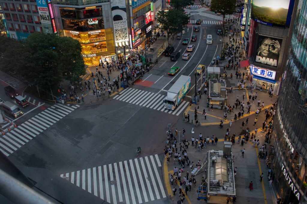 Elevated evening view of Shibuya Crossing in Tokyo with crowds gathering at crosswalks, bright storefronts, and busy city streets.