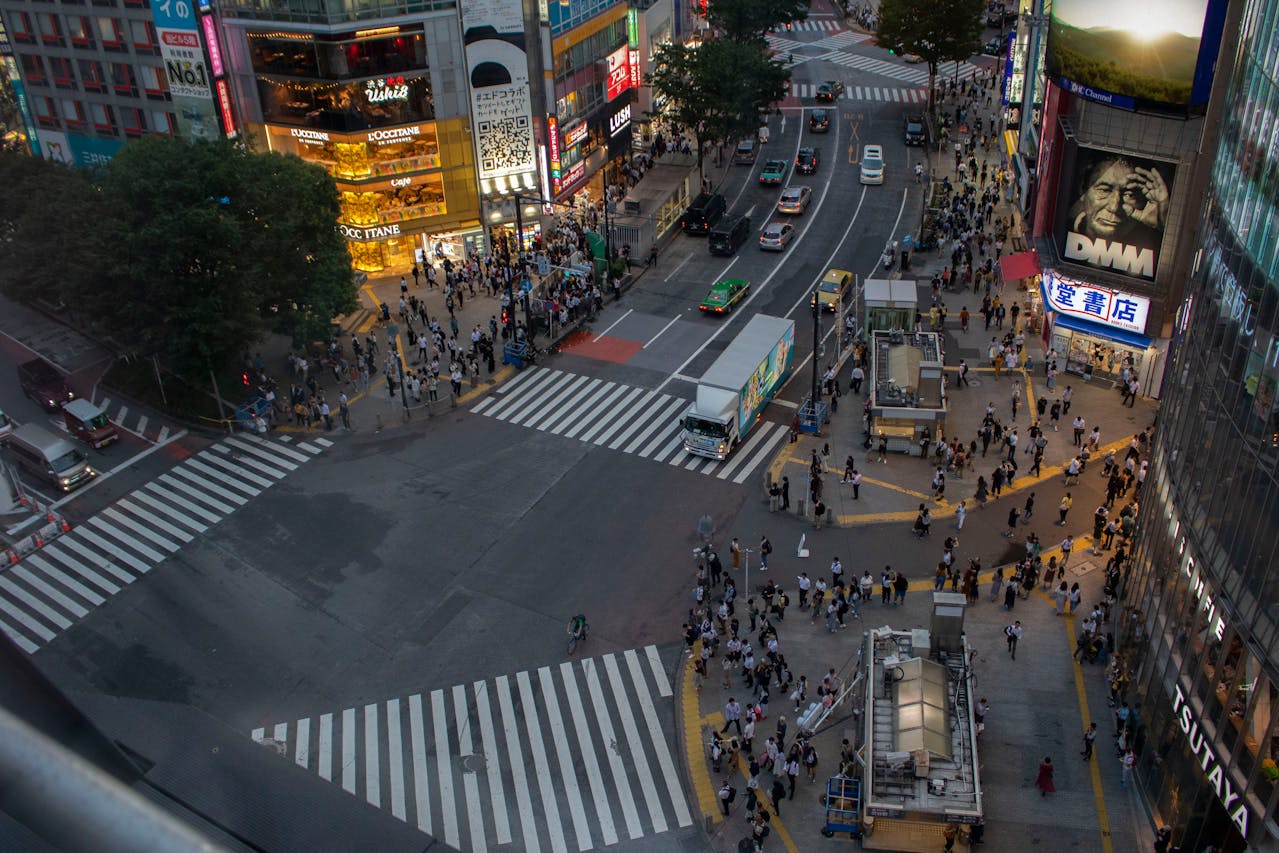 Elevated evening view of Shibuya Crossing in Tokyo with crowds gathering at crosswalks, bright storefronts, and busy city streets.