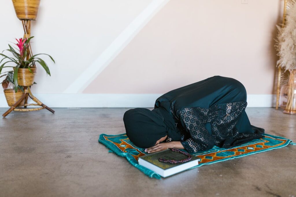 Muslim woman praying on a prayer mat indoors with Quran and prayer beads beside her