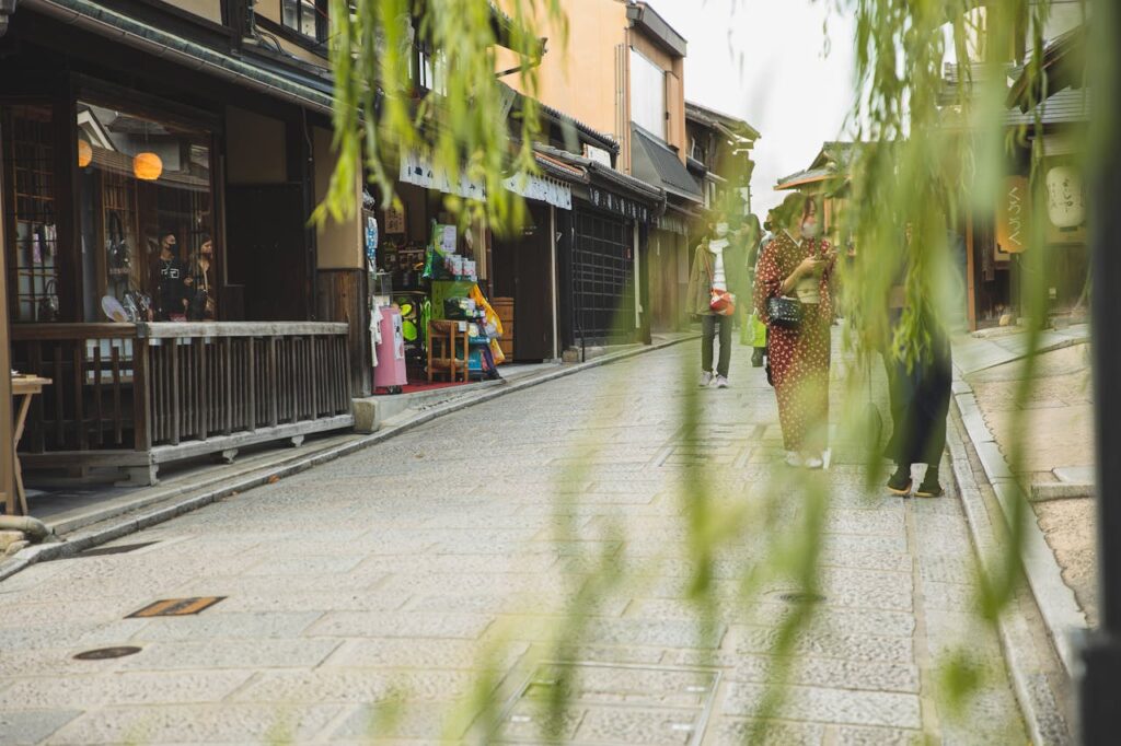Traditional street scene in Kyoto’s Gion district with historic wooden buildings, willow branches in the foreground, and visitors walking in kimono.