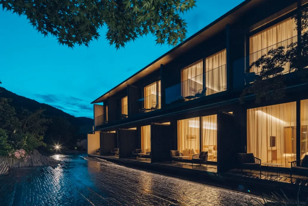 Exterior view of MUNI Kyoto by Onko Chishin at dusk, featuring contemporary architecture, illuminated guest rooms, and a tranquil waterside setting in Arashiyama.