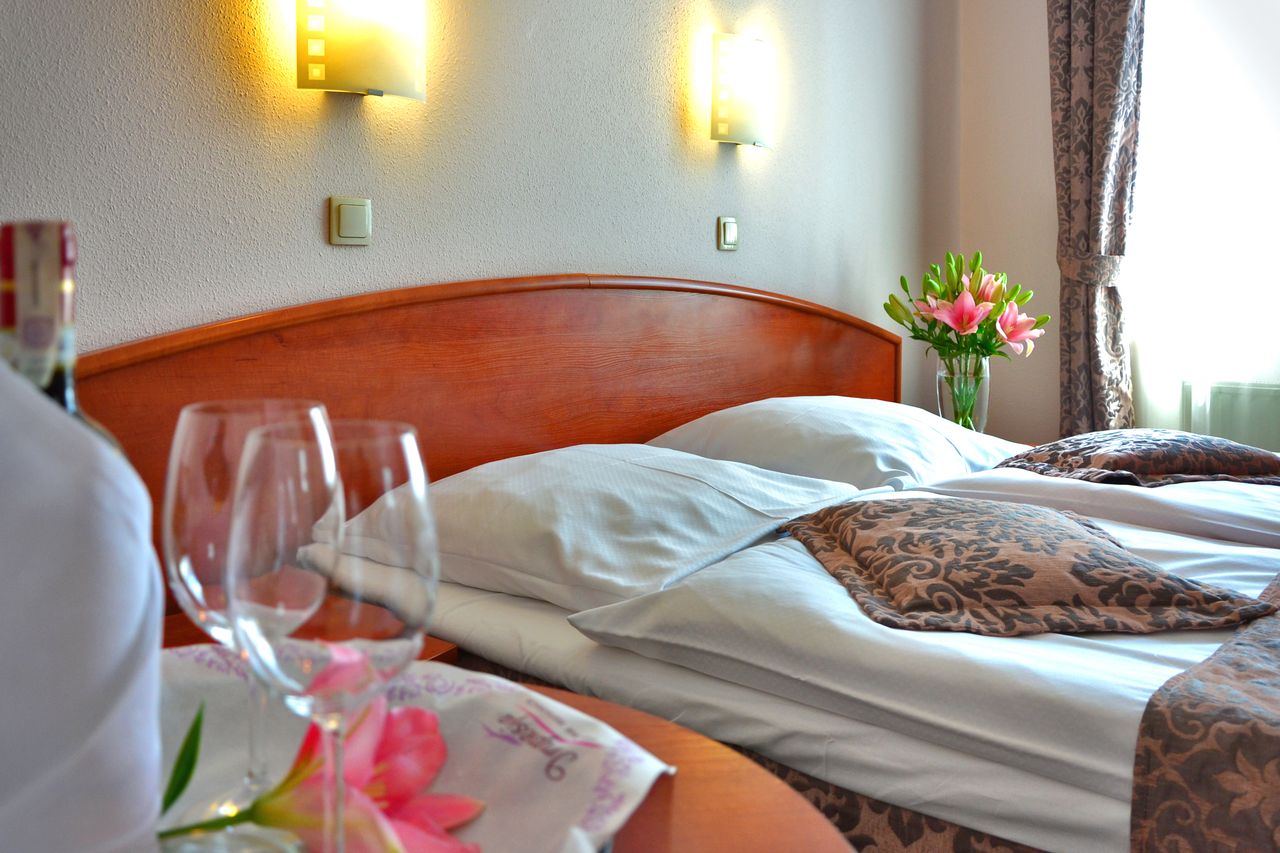 Hotel room with wooden headboard, white pillows, and pink lily flowers on the bedside table with wine glasses in the foreground
